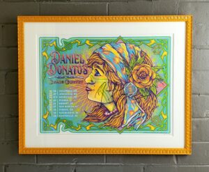 Horizontal Daniel Donatos concert print depicting the profile of a woman with a head scarf/flower. This is colored in greens, blues, yellow, oranges, and burgundy. This is framed in a schoolbus yellow fluted frame with a white mat.