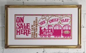 Vintage Pink Floyd advert that says On Sale Here - The New Projected Sound - See Emily Play. The print is in bronze and fuschia and depicts child like drawing of people on a toy train. This is framed in a matching white mat and a scoop panel closed corner 22k gold frame with a carved flower corner detail with lowlights of red clay.