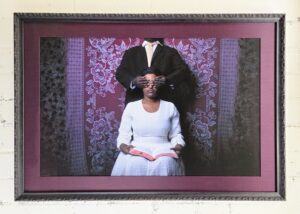 woman in white dress with book in lap has her eyes covered by a mans hands standing behind her. he is dressed in a black suit with a white shirt. His tie is ivory. They are in front of a purple textile. She is dark skinned and the man is even darker skinned. The mat is purple to match the pattern on the textile. The frame is a carved black 'plum' frame