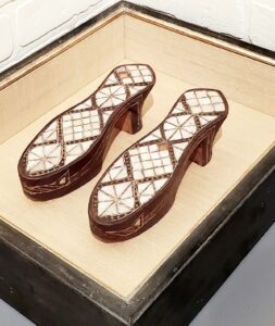 mother of pearl inlaid sandal shoe bases, side by side in warm black shadowbox frame with white gold lip. Box is lined with tan textured silk.